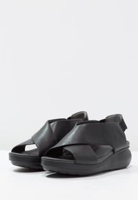 Sandali neri in pelle con design a fascetta incrociata, punta aperta e suola imbottita per il comfort. Presentano un tacco basso e arrotondato e una fascetta regolabile.