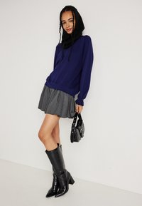 Lila kapucnis pulóver, szürke csíkozott rakott szoknya, fekete térd fölé érő hegyes csizma és egy kis fekete kézitáska.