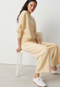 Femme aux longs cheveux foncés assise sur une chaise blanche, portant un sweat-shirt beige et un pantalon large assorti avec des baskets blanches.