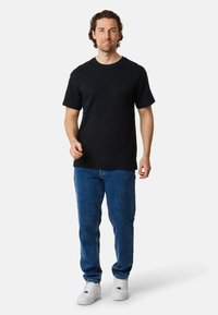 Black short-sleeve t-shirt paired with blue jeans and white sneakers; model stands upright with a neutral expression against a plain background.