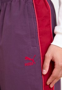 Violet sportiniai džinsai su raudonomis šoninėmis juostomis ir raudonu išsiuvinėtu Puma logotipu. Audinys yra lygus su nedideliu blizgesiu.
