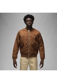 Giacca bomber marrone con zip frontale, polsini e orli a coste, e tasche laterali. Presenta un logo nero sul petto. Tessuto liscio.