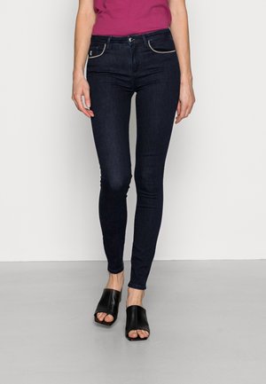 Mujer con vaqueros ajustados azul oscuro, una camiseta magenta y zuecos de tacón abiertos negros, de pie sobre un suelo gris claro.