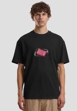Ung mand iført sort T-shirt med et rødt sæbebar-grafik mærket "FIGHT CLUB" og beige bukser, stående mod en ensfarvet baggrund.