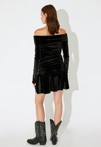 Vestido negro de terciopelo sin hombros, con mangas largas, cuerpo fruncido y dobladillo acampanado, combinado con botas de vaquero negras con bordados.