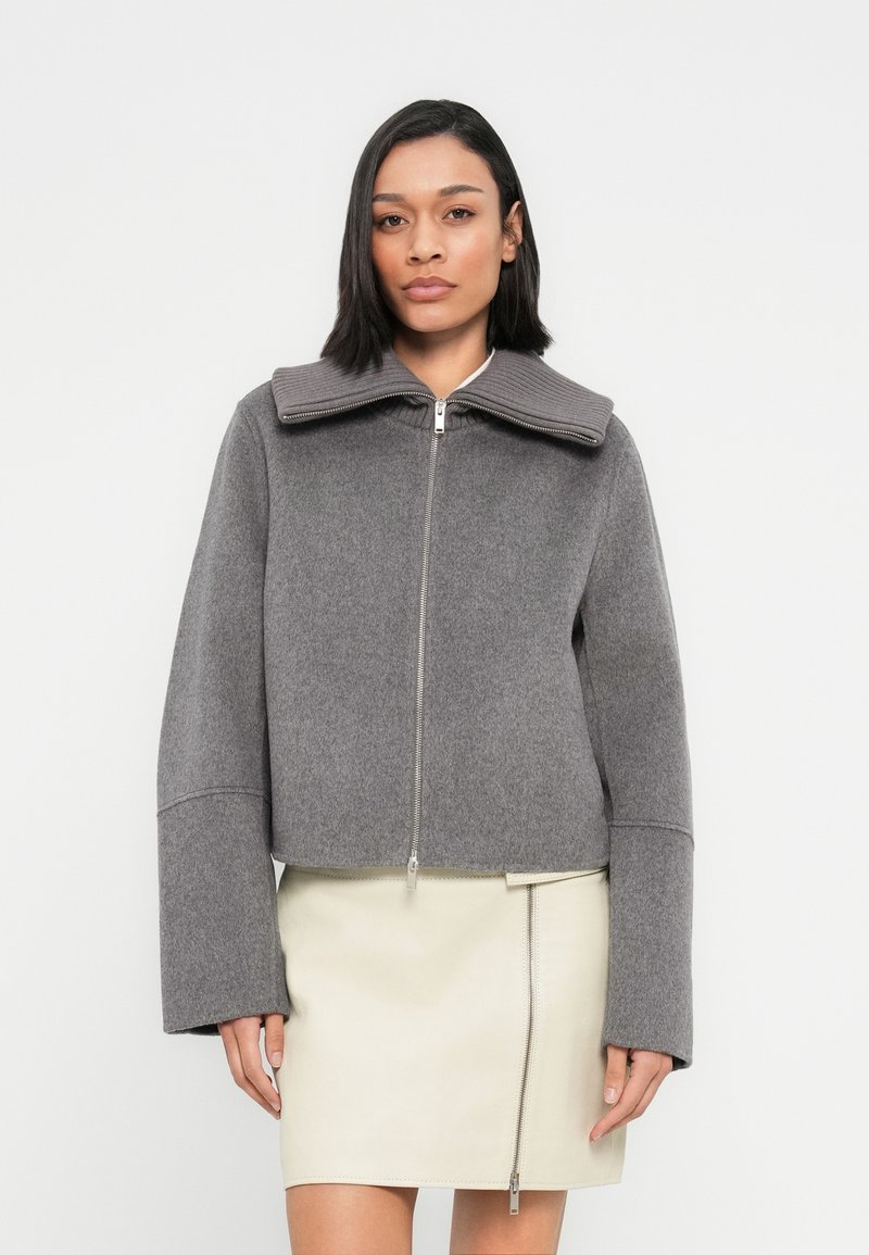 Γκρι κοντό μπουφάν με φαρδιά ριμπ ύφανση στο γιακά, κλείσιμο με φερμουάρ μπροστά και φαρδιά μανίκια, συνδυασμένο με κρέμ δερμάτινη φούστα.