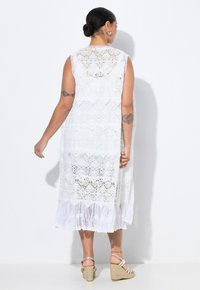 Abito bianco in pizzo con design trasparente, senza maniche e orlo a volant. Presenta motivi intricati e una silhouette fluida.