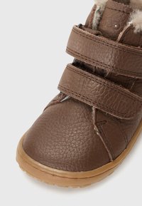 Bruine leren schoen met een gestructureerde afwerking, voorzien van twee klittenbandsluitingen en een rubberen zool. Warme voering zichtbaar aan de bovenkant.