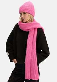 Różowy prążkowany dziergany komin i pasująca czapka wykonane z miękkiego materiału, zestawione z ciemnym, oversize'owym swetrem. Szalik delikatnie opada wokół szyi.