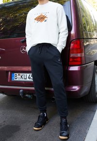 Bílé mikina s logem "Air Jordan" v oranžové a černé barvě; černé tepláky s logem Jordan; černé tenisky s gumovou podrážkou.