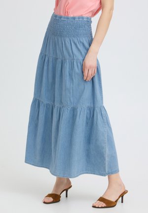 Femme portant une jupe maxi à volants bleu clair avec une taille smockée, un haut rose et des mules à talons ouvertes marron, debout devant un fond uni.