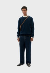 Pull en polaire bleu marine avec des poignets côtelés, porté par-dessus une chemise blanche. Pantalons en denim foncé à coupe ample et chaussures sombres. Sac bandoulière marron.
