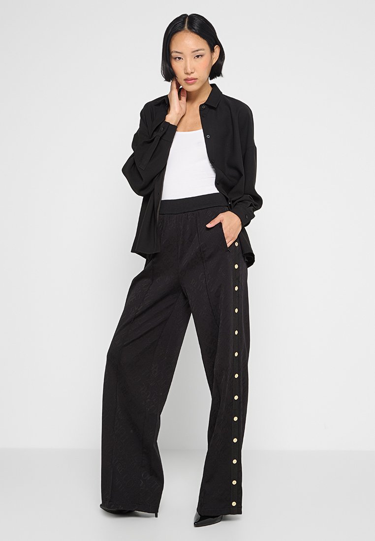 Schwarzes Hemd mit Knöpfen über einem weißen Tank-Top, kombiniert mit schwarzen, hochtaillierten, weit geschnittenen Hosen, die goldene Druckknöpfe an der Seite haben.