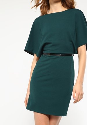 Kobieta w ciemnozielonej sukience do kolan z szerokimi krótkimi rękawami i cienkim czarnym paskiem w talii, stojąca na białym tle.