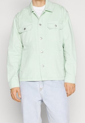 Mann trägt eine hellgrüne, zum Knöpfen vorgesehene Jacke mit Klappentaschen auf der Brust über einem weißen Hemd und hellblauen Jeans, steht vor einem schlichten Hintergrund.