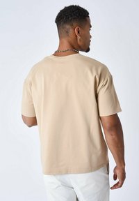 Camiseta beige de manga corta con un corte relajado, textura suave, cuello redondo y aberturas laterales, mostrada desde la parte trasera con detalles de costura visibles.