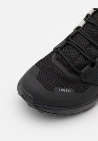 Zapatilla deportiva negra con parte superior de malla transpirable, superposiciones texturizadas y suela de goma. Presenta cordones elásticos y etiqueta "RAIN.RDY".