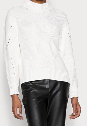 Weißer Strickpullover mit Rollkragen und Zopfmuster, mit gerippten Bündchen und Saum, kombiniert mit schwarzen Lederhosen an einer stehenden Person.