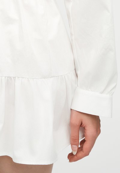 Λευκό φόρεμα με μακριά μανίκια και λεπτομέρεια σούρωσης στη μέση, μαζί με μια κοντινή λήψη ενός χαλαρού χεριού δίπλα στο τελείωμα.