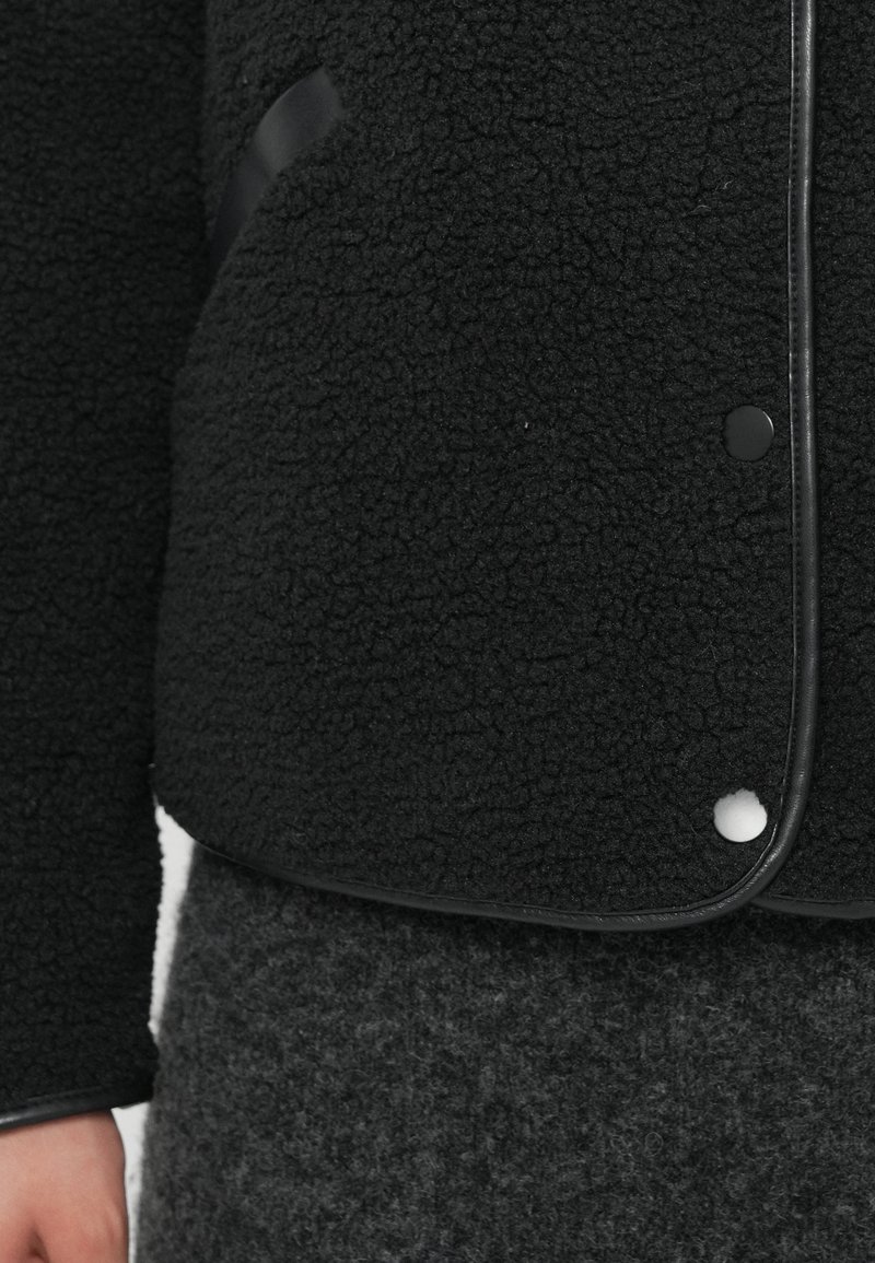 Veste en polaire noire texturée avec des boutons ronds et des finitions en cuir, associée à un pantalon en laine gris foncé, montrant la partie médiane et la main gauche.