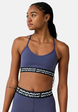 Jonge vrouw met gevlochten haar die een paarse sport-bh en legging set draagt met zwarte elastische banden met het label "Brooks."