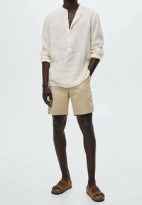 Camicia di lino beige chiaro con colletto parziale e bottoni, abbinata a pantaloni corti beige chiaro e sandali marroni. Design semplice, vestibilità rilassata.