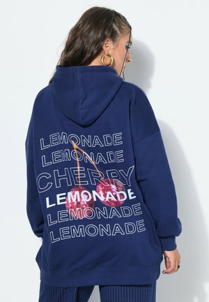 Kvinde iført navyblå hættetrøje med gentaget hvid tekst "LEMONADE," "CHERRY," og to store røde kirsebær trykt på bagsiden.