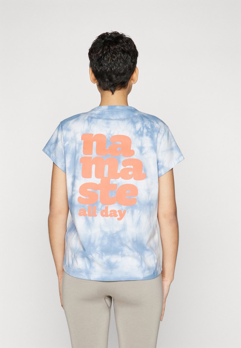 Sīkpēdaina īsām piedurknēm autentiskā tīņu stila T-kreklā gaiši zilā krāsā ar lielu oranžu grafiku uzrakstu "namaste all day" aizmugurē. Mīksta kokvilnas auduma.