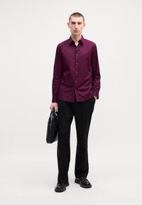 Burgunderfarget langermet skjorte med klassisk krage, knappestolpe og glatt stoff. Bæres med svarte bukser og holder en svart skinnveske.