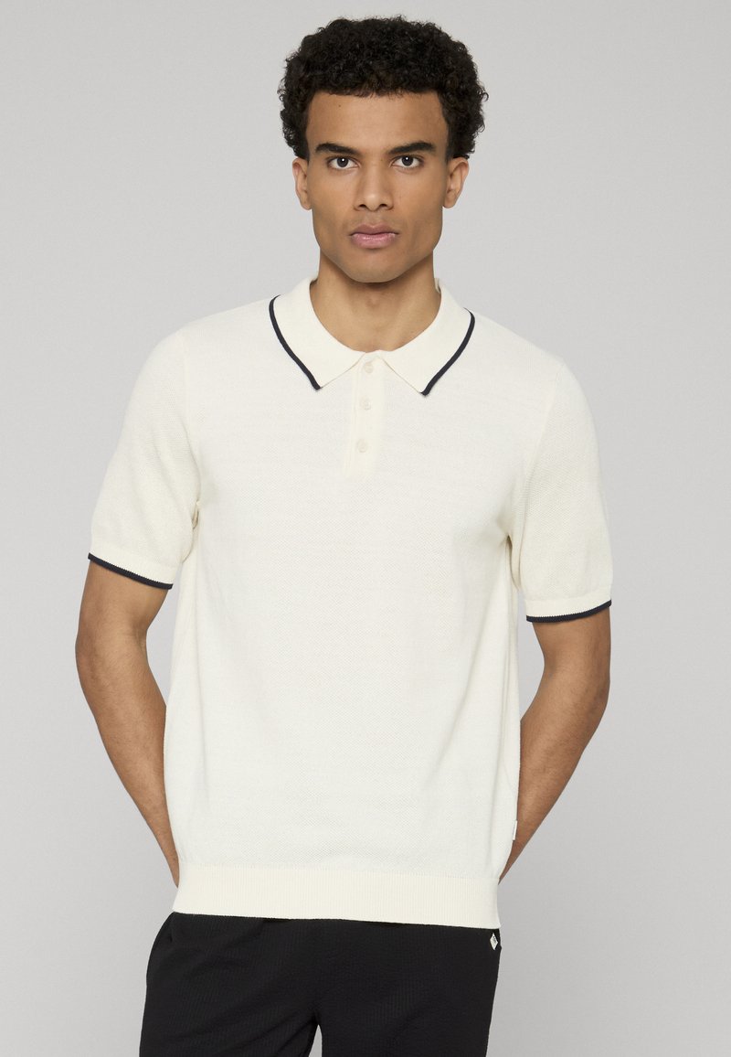 Polo crème en maille texturée, manches courtes, patte de boutonnage à trois boutons, et bordure marine au col et aux manches. Design décontracté.