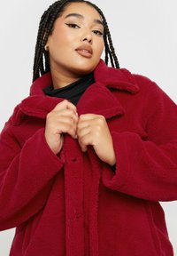 Cappotto in fleece bordeaux con colletto ampio e chiusura con bottoni, caratterizzato da una texture morbida e grandi tasche frontali.