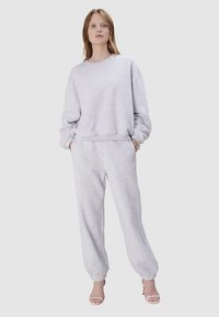 Sweat-shirt en coton gris et pantalon de survêtement assorti avec poignets élastiques, coupe ample et col rond. Le modèle est debout, les mains dans les poches.