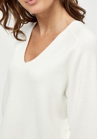 White, soft knit sweater with a deep V-neckline and relaxed fit. Notable features include textured fabric and drop shoulder design.