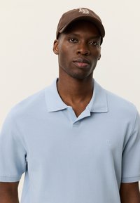 Ανοιχτό μπλε πόλο μπλουζάκι με διακριτικό υφασμάτινο μοτίβο και μικρό κεντημένο έμβλημα. Συνδυάζεται με καφέ καπέλο με λογότυπο.