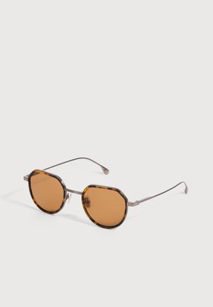 Lunettes de soleil octogonales en écaille de tortue avec des branches fines en métal argenté et des verres teintés ambre sur une surface blanche.