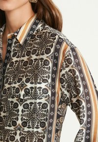 Hemd in Creme mit schwarzen Blumenmustern, braunen und orangefarbenen Akzentstreifen sowie schwarzen Knöpfen an der Fronttasche. Weicher Stoff mit angenehmer Textur.