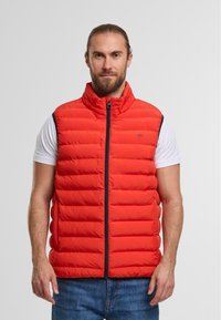 Rote Pufferweste mit hohem Kragen, Frontreißverschluss und horizontalen Steppnähten. Über einem weißen Hemd getragen, kombiniert mit blauen Jeans.