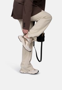 Beige sportieve schoenen met luipaardprintaccenten, gemêleerde panelen en witte veters. Draagt een lichte broek en een bruine overshirt.