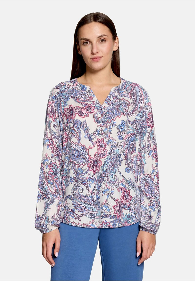 Femme portant un chemisier à manches longues avec un motif floral paisley rose et bleu et un pantalon bleu, faisant face à l’avant avec une expression neutre.