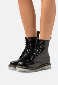 Glänsande svarta lackskinnsskor med snörning och gul söm samt genomskinliga sulor, bärs på bara ben mot en vit bakgrund.