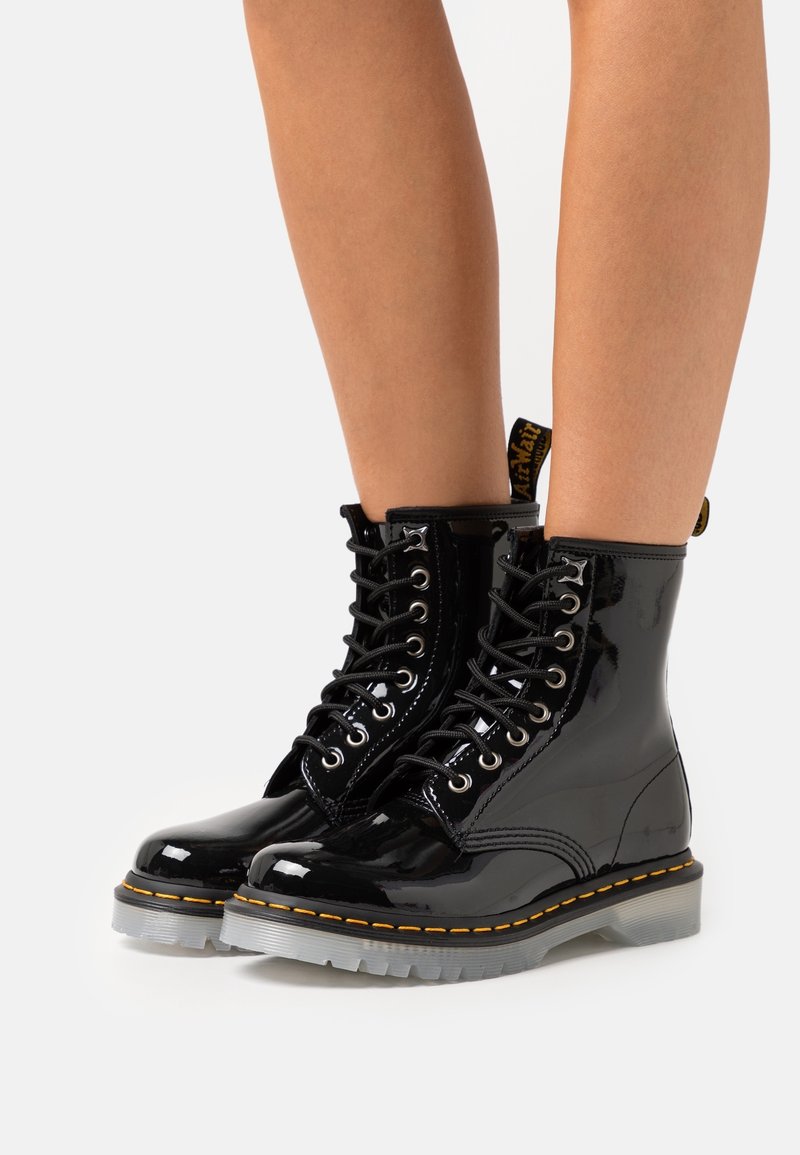 Glänsande svarta lackskinnsskor med snörning och gul söm samt genomskinliga sulor, bärs på bara ben mot en vit bakgrund.