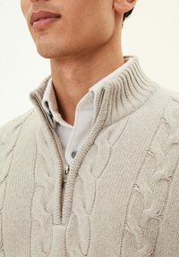 Beige cable-knit sweater with a high ribbed collar and silver zipper, layered over a white collared shirt. Textured knit pattern is prominent.