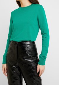 Grön långärmad tröja kombinerad med högmidjade svarta byxor i konstläder med en texturerad krokodilform och en slät yta.