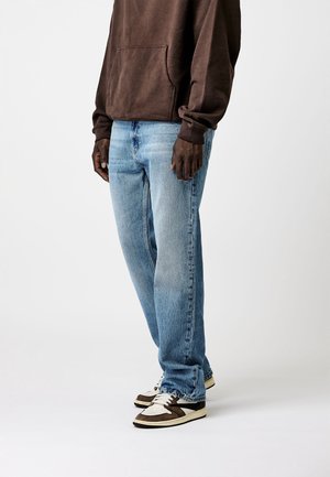 Homme portant un jean droit bleu clair, un sweat à capuche marron et des baskets blanches et marron, debout devant un fond uni.