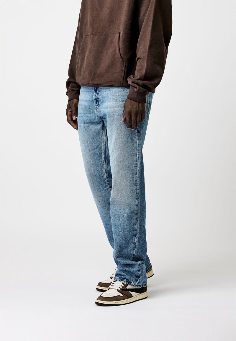 Homme portant un jean droit bleu clair, un sweat à capuche marron et des baskets blanches et marron, debout devant un fond uni.
