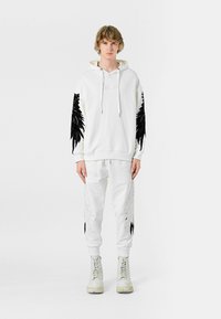 Sudadera blanca con diseños negros parecidos a plumas en las mangas, combinada con pantalones blancos con patrones similares. Llevada con botas de color claro.