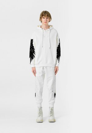 Sudadera blanca con diseños negros parecidos a plumas en las mangas, combinada con pantalones blancos con patrones similares. Llevada con botas de color claro.
