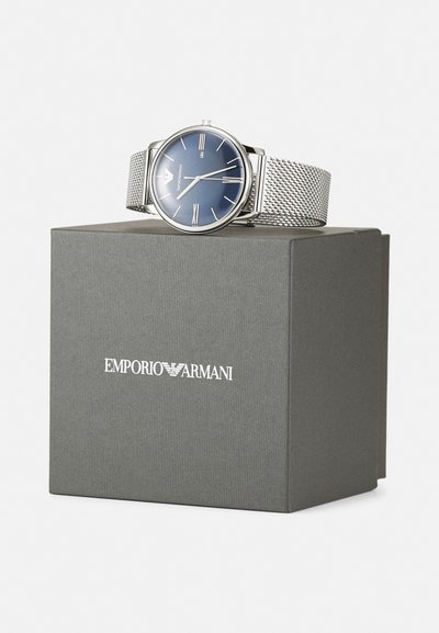 Silbernes Netzarmbanduhr mit blauem Zifferblatt und silbernen römischen Ziffern als Stundenmarkierungen, präsentiert auf einer grauen Box mit dem Aufdruck "Emporio Armani".