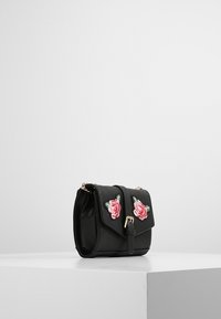 Borsa nera in ecopelle con chiusura a cappottina, caratterizzata da dettagli ricamati di rose rosa e una tracolla in metallo dorato. Forma compatta e rettangolare.