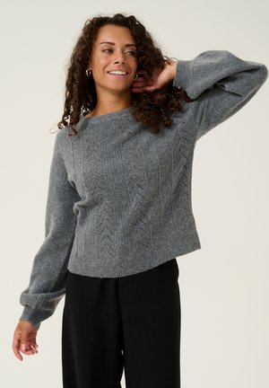 Grauer gestrickter Pullover mit einem strukturierten Zickzackmuster, weiten Ärmeln und einer verkürzten Passform, kombiniert mit schwarzen Hosen.